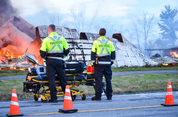 Two paramedics stand by a stretcher near the burning wreckage of a crashed aircraft during emergency response.